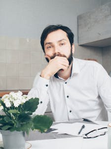 A man in a white shirt thinking, seated at a desk with papers and a plant.