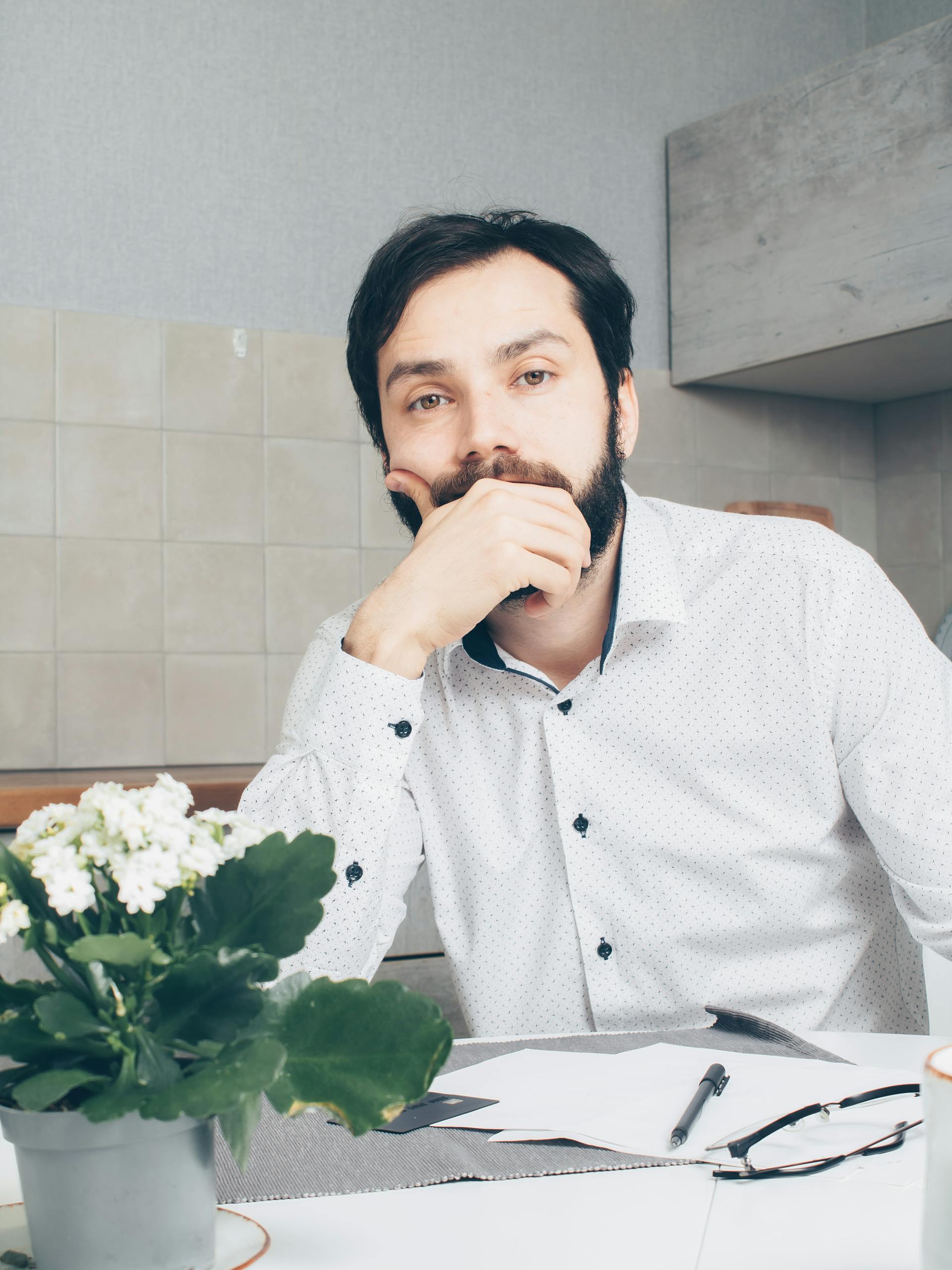 A man in a white shirt thinking, seated at a desk with papers and a plant.