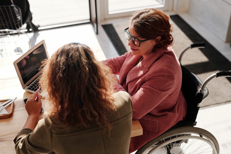 Two women working on laptops, one using a wheelchair, collaborating in a modern café.